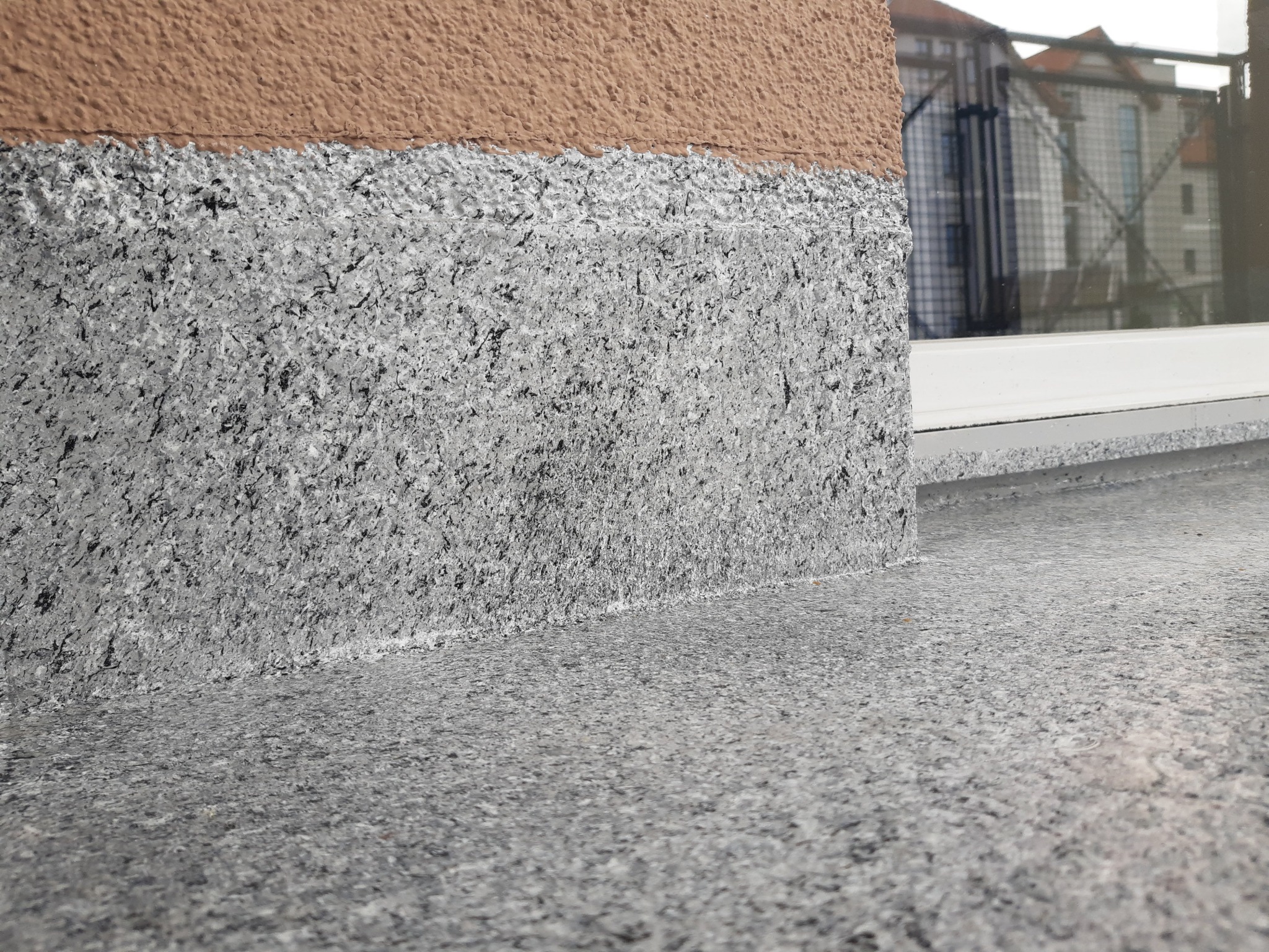 Wykończony fragment balkonu z szarą, kamienną posadzką i fragmentem elewacji w kolorze brązowym, widok z niskiej perspektywy.