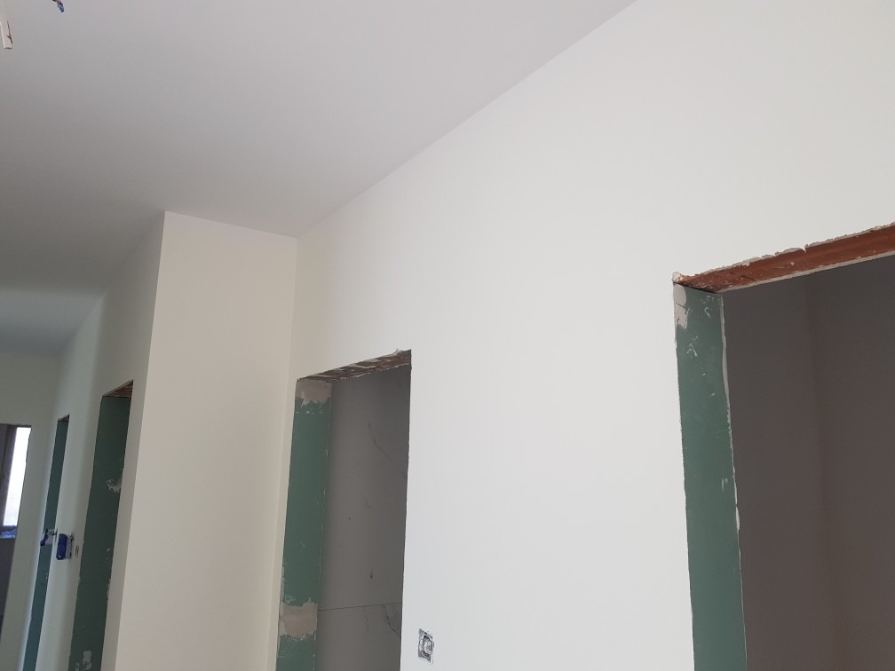 Świeżo pomalowane, jasne wnętrze z ościeżnicami drzwiowymi przygotowanymi do montażu, widoczne niedoskonałości w wykończeniu wokół otworów drzwiowych i gniazdko elektryczne na ścianie.