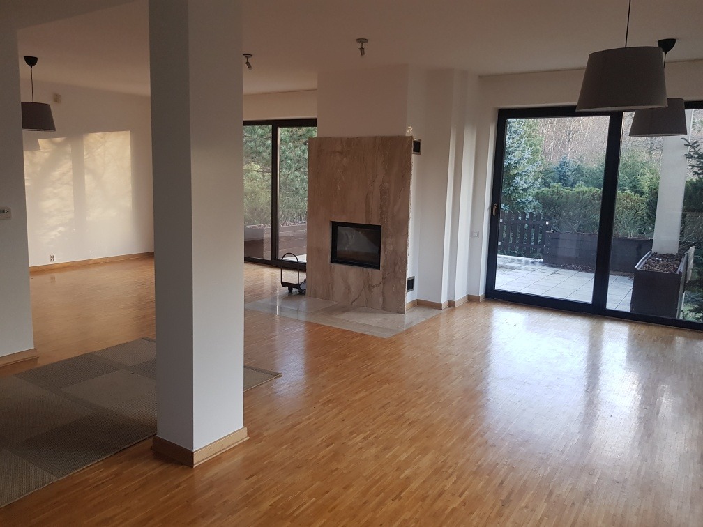 Jasne, przestronne wnętrze z parkietem, marmurowym kominkiem i dużymi oknami tarasowymi wychodzącymi na zieleń. Ściany świeżo pomalowane na biało, widoczny fragment kolumny.
