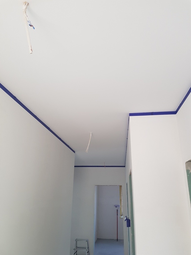 Przygotowanie do prac wykończeniowych: pomieszczenie z zabezpieczonymi taśmą malarską krawędziami ścian i sufitu, widoczna drabinka i wałek do nakładania farby w tle.