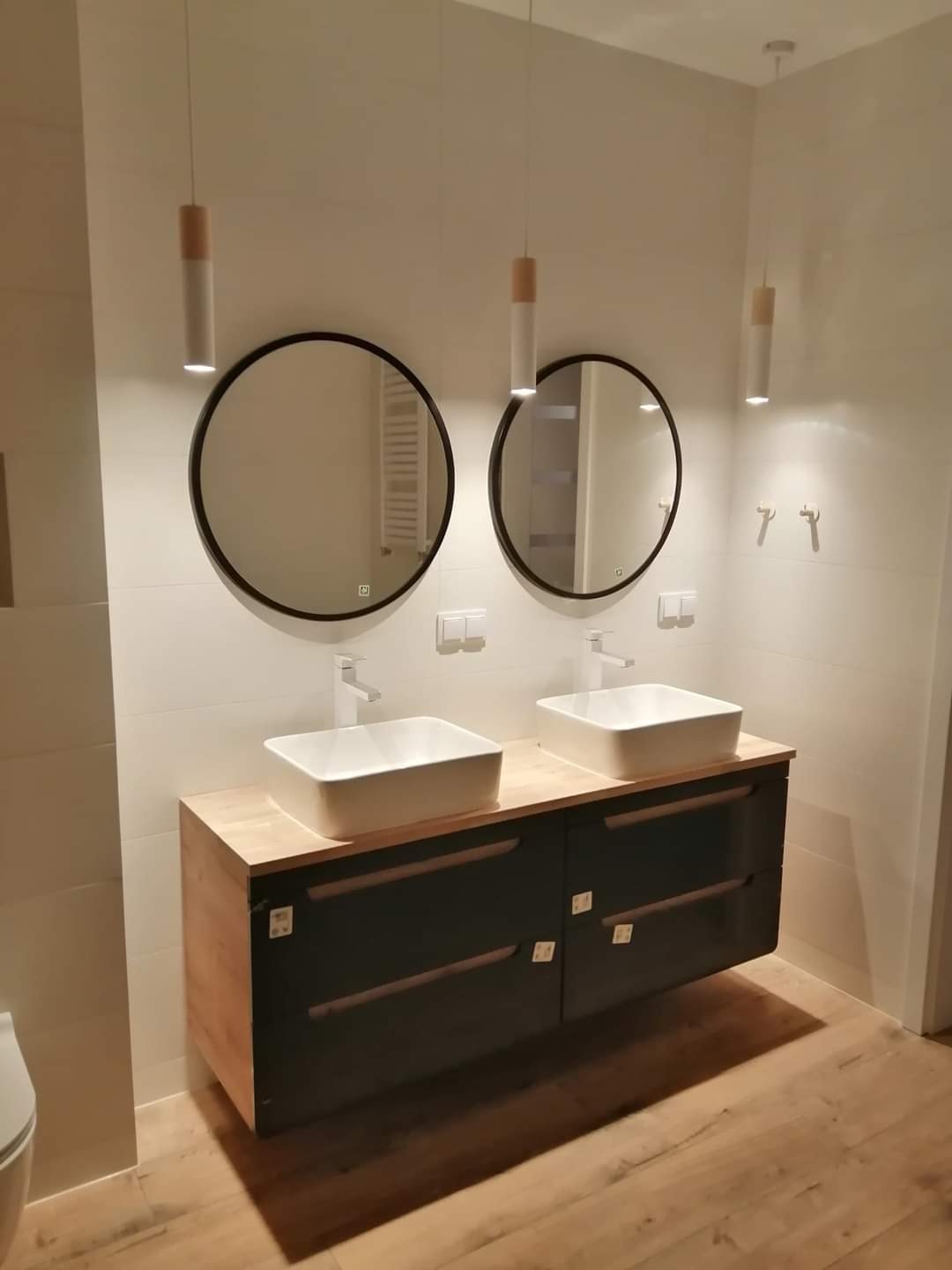 Nowoczesna łazienka z dwoma kwadratowymi umywalkami nablatowymi, okrągłymi lustrami w czarnych ramach i wiszącą szafką z drewnianym blatem i ciemnymi frontami, oświetlona wiszącymi lampami...