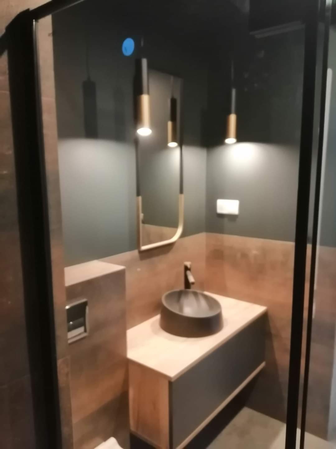 Nowoczesna łazienka z umywalką nablatową w odcieniach szarości i brązu, oświetlona wiszącymi lampami z elementami złotymi i lustrem w cienkiej złotej ramie.