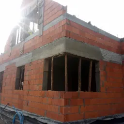 Surowy stan budynku z czerwonej cegły, widoczne otwory okienne z drewnianymi stemplami podtrzymującymi nadproża, oraz pracownik w oknie na poddaszu.