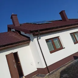 Nowoczesny dom z białą elewacją, brązowym dachem z panelami słonecznymi, brązowymi oknami i otwartymi drzwiami wejściowymi, sfotografowany w słoneczny dzień.