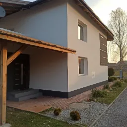 Nowoczesny dom z jasnoszarą elewacją, drewnianym zadaszeniem nad wejściem i fragmentem ocieplenia na bocznej ścianie, widok z poziomu gruntu.