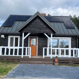 Elewarm - Czarny dom z drewnianą elewacją, białymi ramami okiennymi i balustradą werandy, z panelami słonecznymi na szarym dachu i nową kostką brukową przed wejściem.