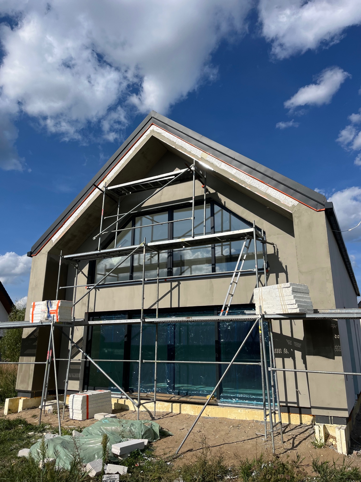 Nowoczesny dom jednorodzinny w trakcie prac elewacyjnych, widoczne rusztowania, przygotowane płyty izolacyjne i świeżo nałożona warstwa tynku w kolorze beżowym, błękitne niebo z chmurami w tle.