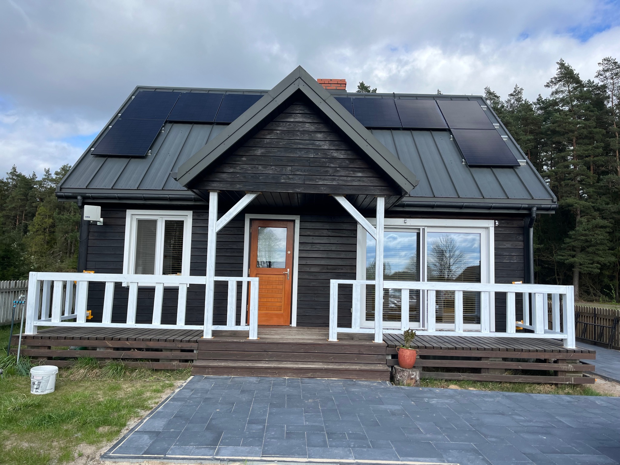 Czarny dom z drewnianą elewacją, białymi ramami okiennymi i balustradą werandy, z panelami słonecznymi na szarym dachu i nową kostką brukową przed wejściem.