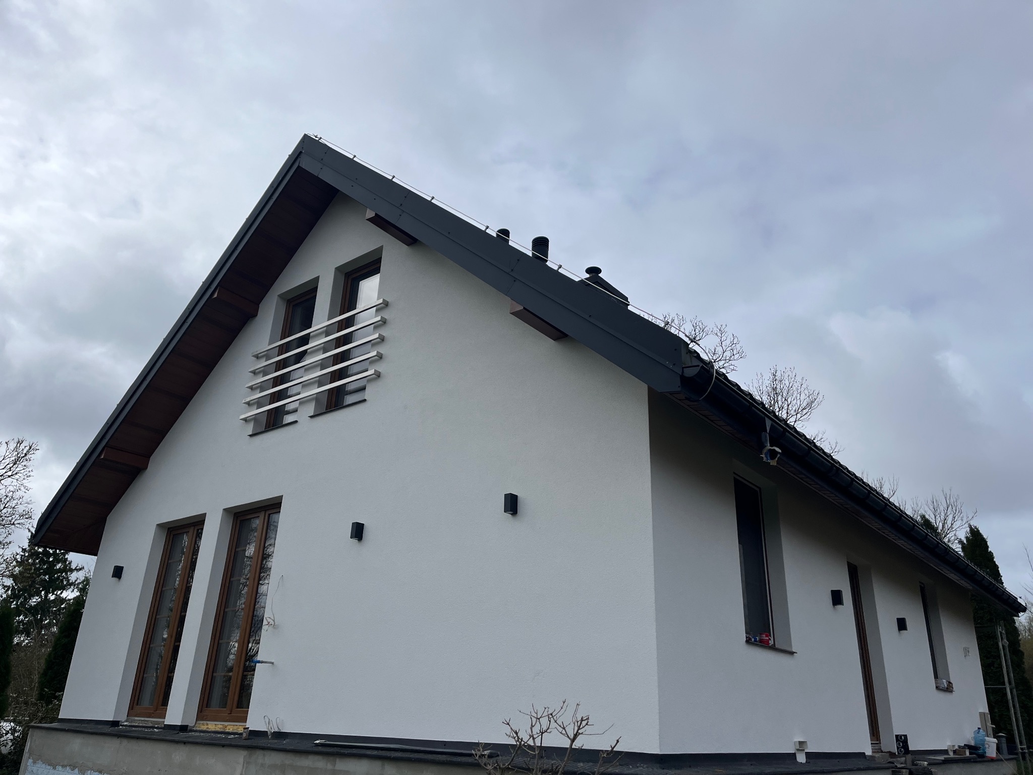 Nowoczesny dom jednorodzinny z białą elewacją, ciemnym dachem i brązowymi ramami okiennymi, widziany pod kątem z dołu w pochmurny dzień.