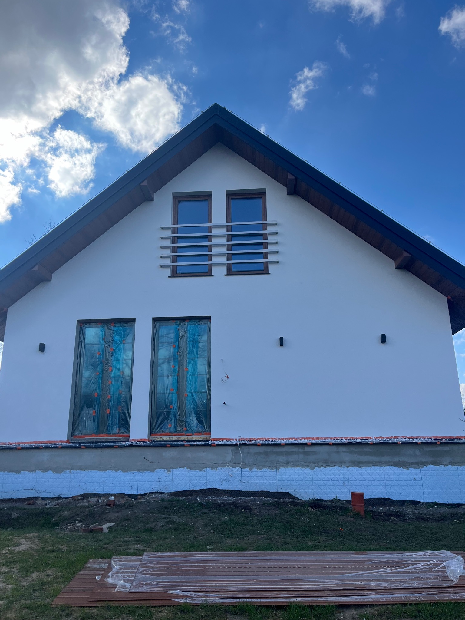 Dom jednorodzinny z białą elewacją, okna zabezpieczone folią malarską, widoczna warstwa izolacyjna przy gruncie i przygotowane deski tarasowe na trawniku.