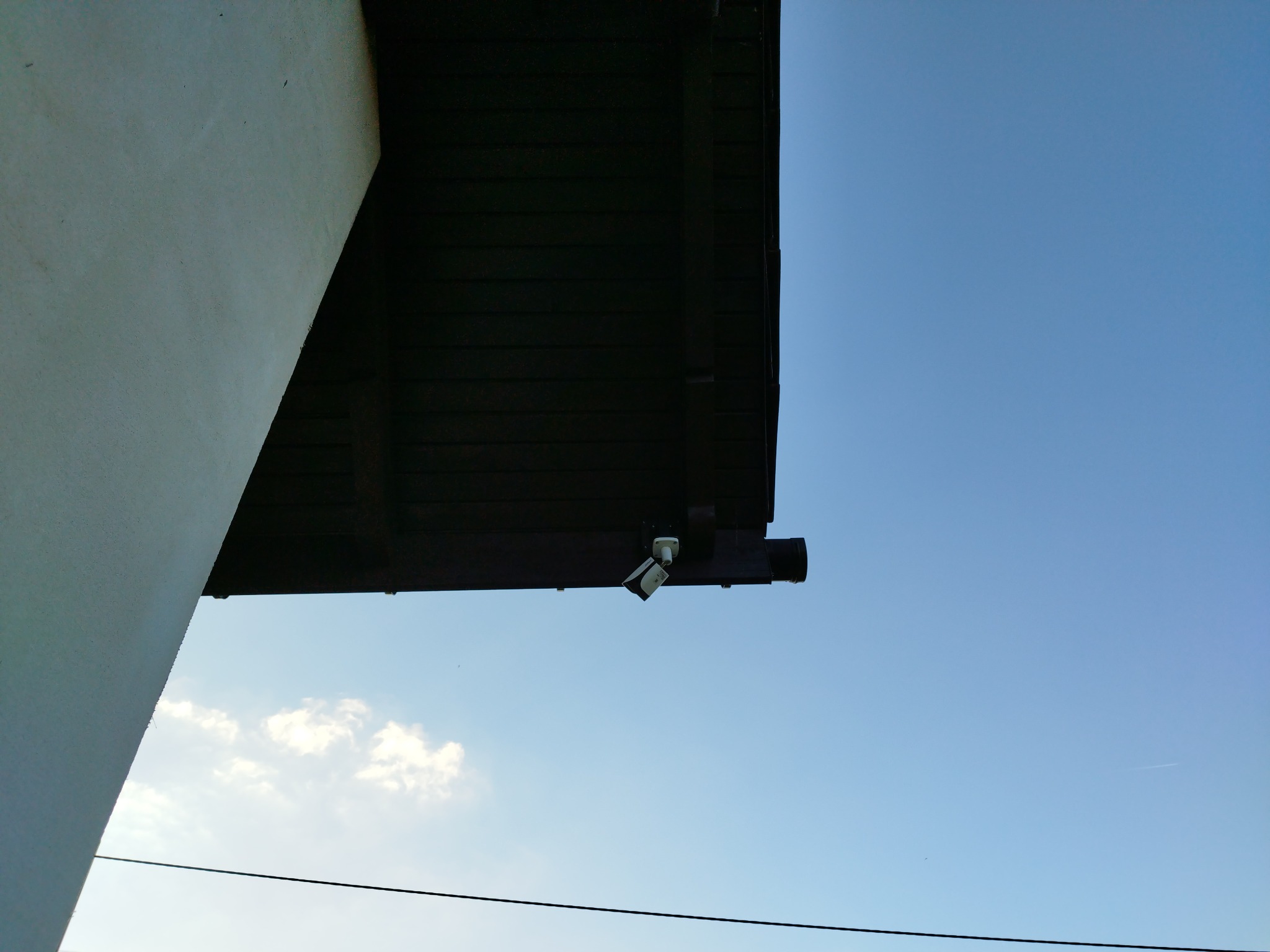 Kamera monitoringu zamontowana pod okapem budynku, widoczna z dołu na tle błękitnego nieba.