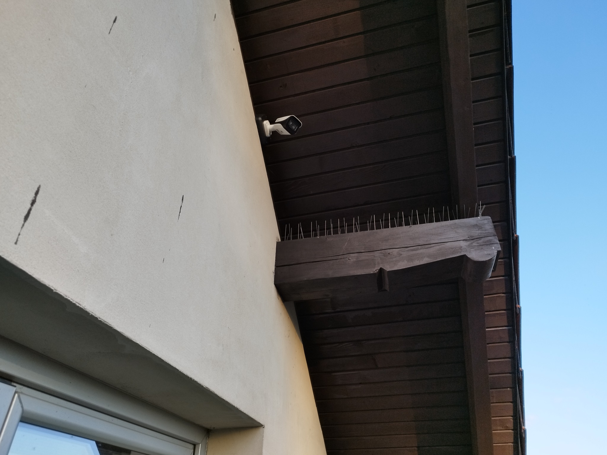 Zewnętrzna kamera bezpieczeństwa zamontowana pod drewnianym okapem z kolcami przeciw ptakom, widok z dołu na jasnym tle nieba.