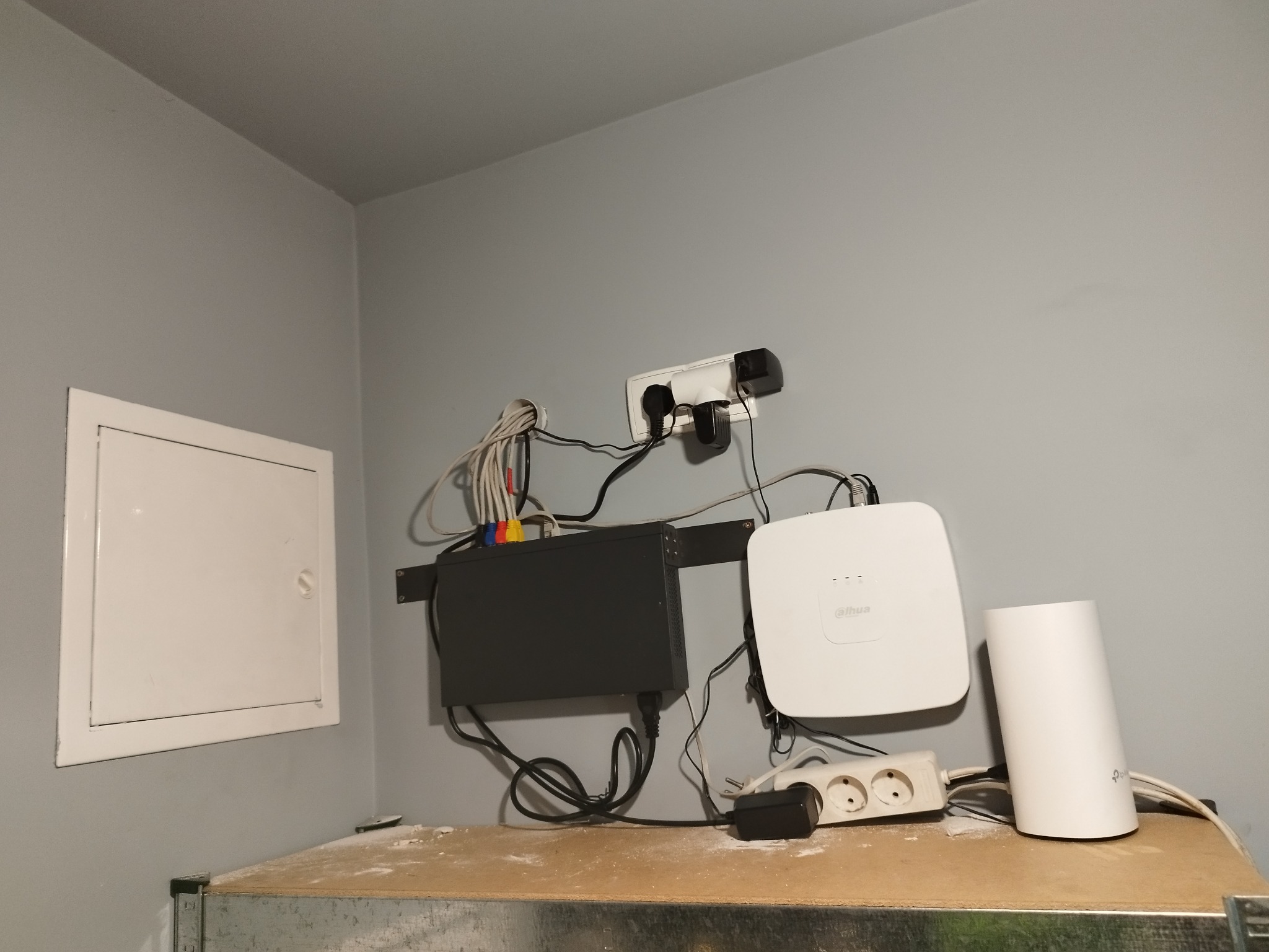 Zamontowany system rejestracji w pomieszczeniu z szarymi ścianami, widoczny rejestrator, biały punkt dostępowy i router, kable zasilające i sieciowe podłączone do gniazdka.