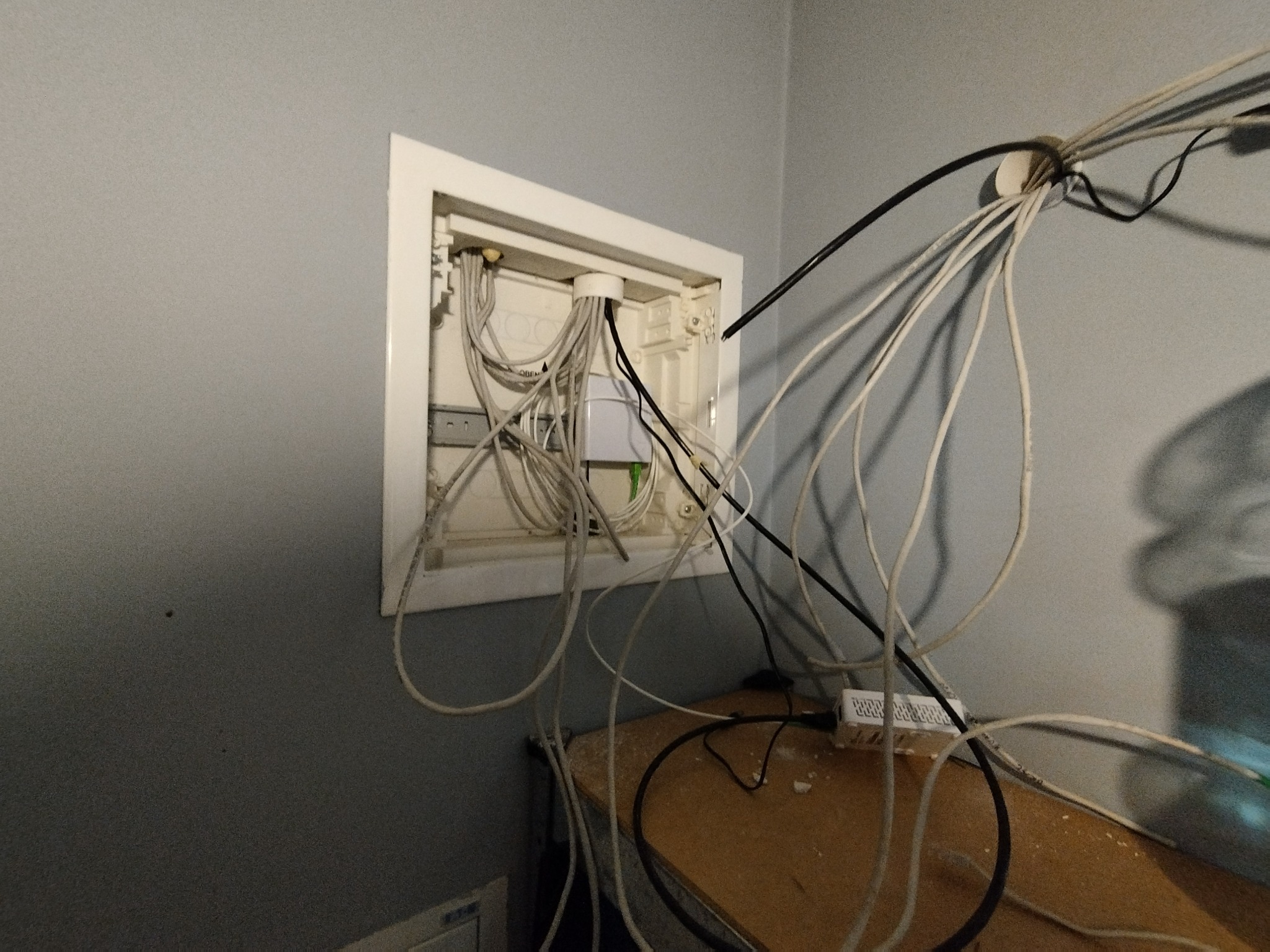 Otwarta skrzynka instalacyjna z licznymi kablami wychodzącymi na zewnątrz, widoczne elementy instalacji wewnątrz oraz router na półce pod spodem, szara ściana w tle.