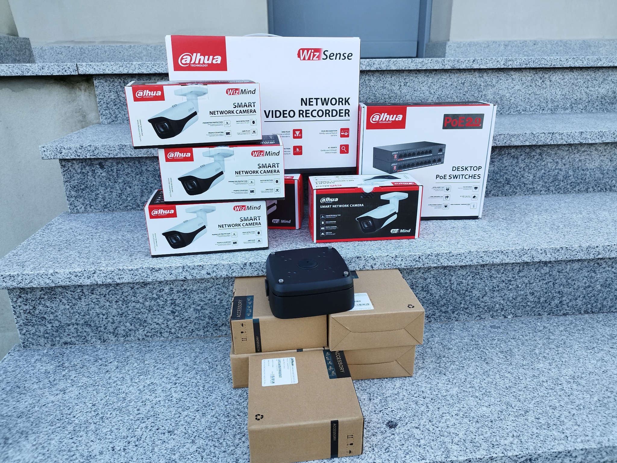 Stos pudełek z kamerami sieciowymi, rejestratorem wideo i przełącznikami PoE marki Dahua, ułożonych na schodach z szarego granitu.