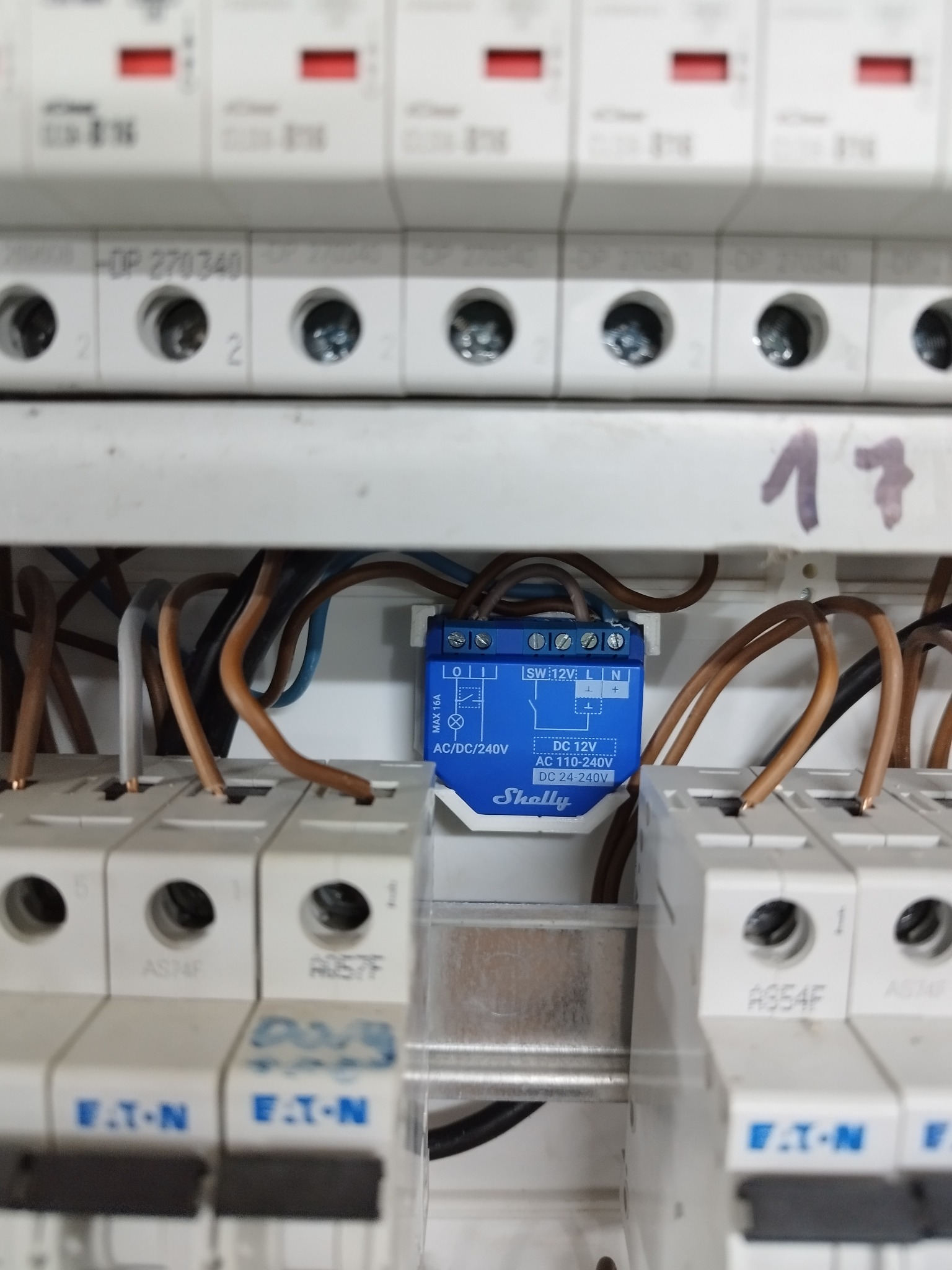 Niebieski moduł Shelly podłączony do przewodów w skrzynce rozdzielczej z bezpiecznikami Eaton, widoczne oznaczenia na module i bezpiecznikach.