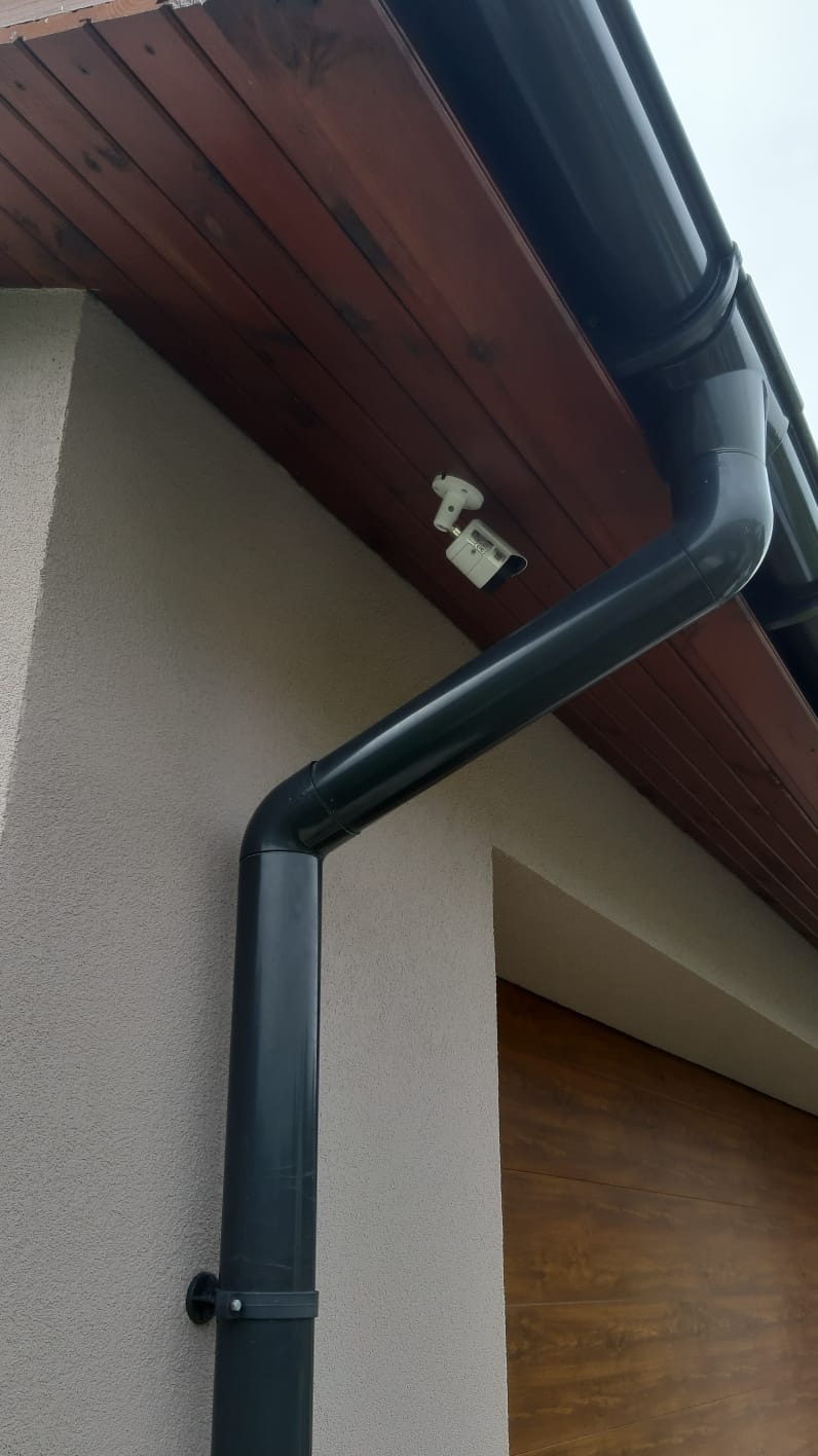 Dwie kamery przemysłowe zamontowane pod drewnianym okapem, obok rynny spustowej na elewacji budynku.