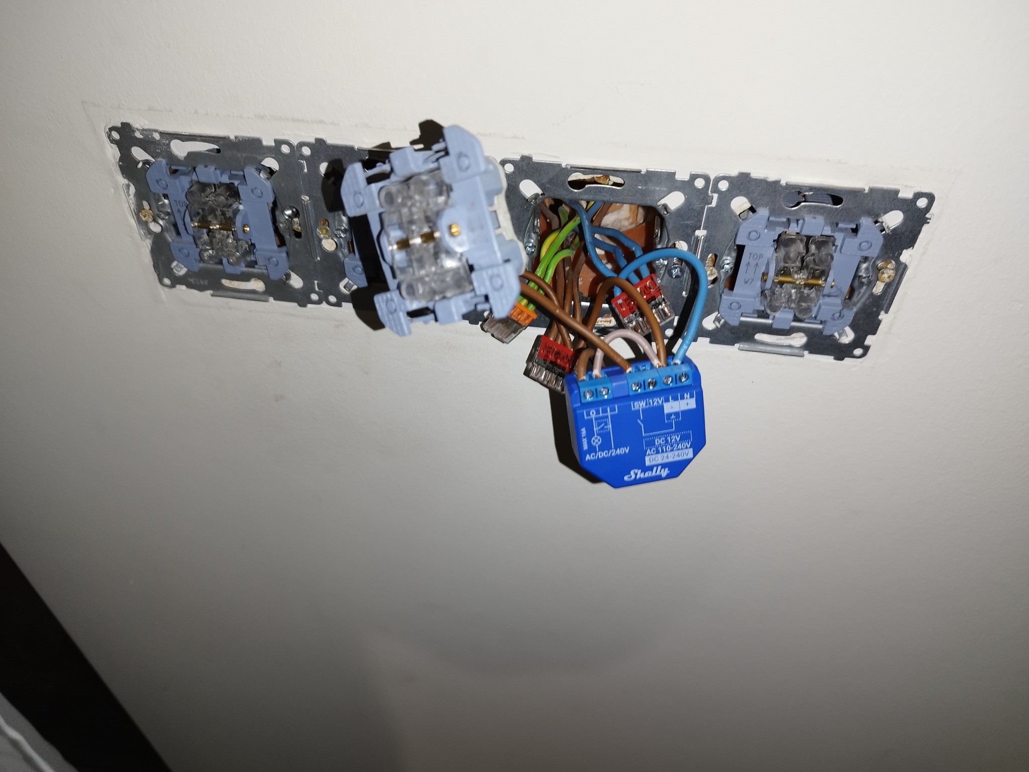 Instalacja inteligentnego włącznika światła Shelly w puszce podtynkowej z widocznymi przewodami i elementami montażowymi, w trakcie podłączenia do istniejącej instalacji elektrycznej.