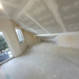 Poddasze po montażu płyt gipsowo-kartonowych i wstępnym szpachlowaniu, widoczne okno dachowe i drzwi balkonowe, ściany przygotowane do dalszych prac wykończeniowych.