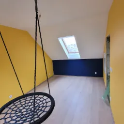 Pomalowane na żółto i granatowo poddasze z oknem dachowym i wiszącym fotelem-huśtawką z czarnego sznura, jasne panele podłogowe.