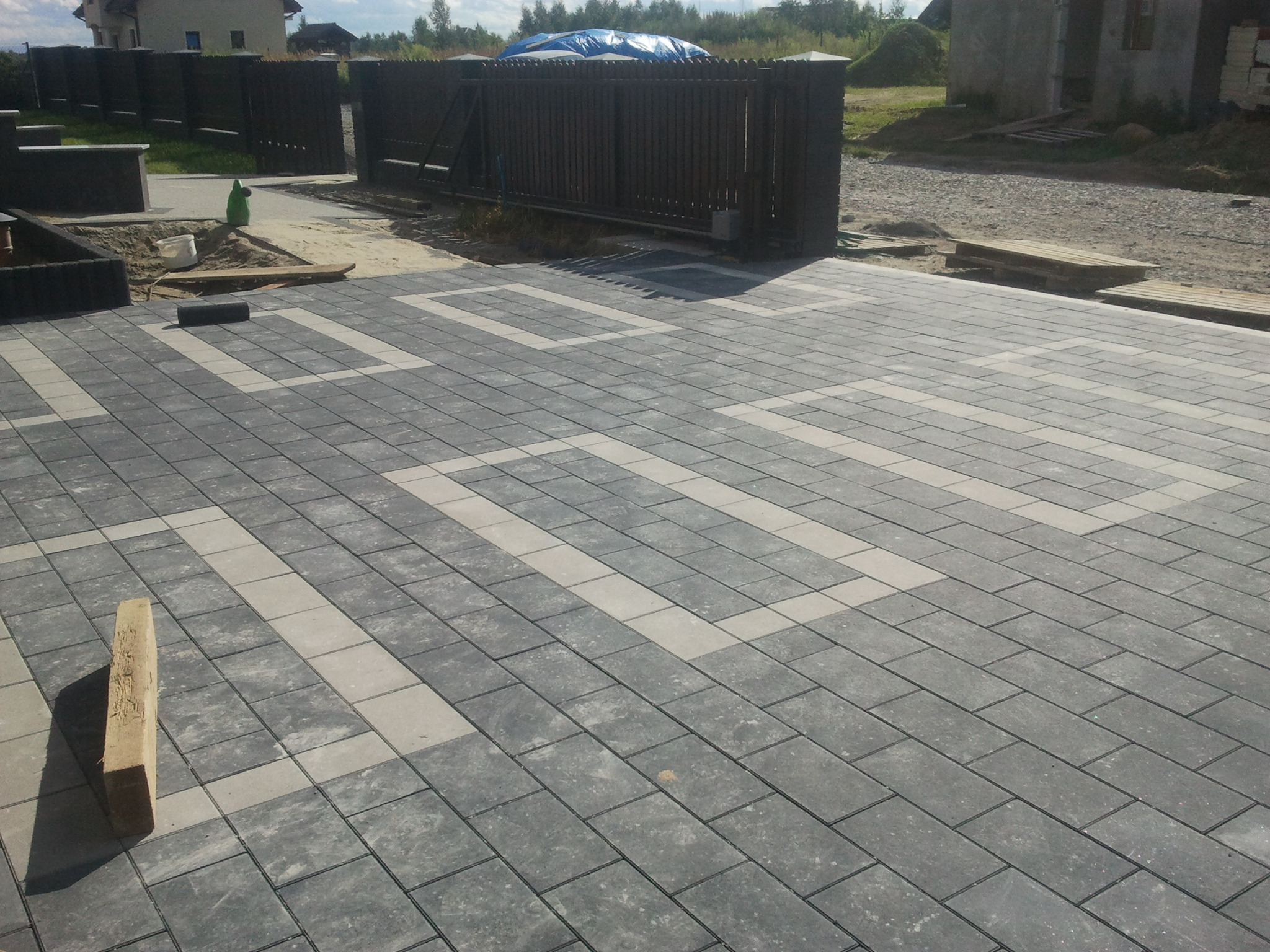Ułożona nawierzchnia z prostokątnej kostki brukowej w odcieniach szarości z jasnymi wstawkami tworzącymi wzór, widoczne przygotowania do dalszych prac brukarskich i ogrodzenie w tle.
