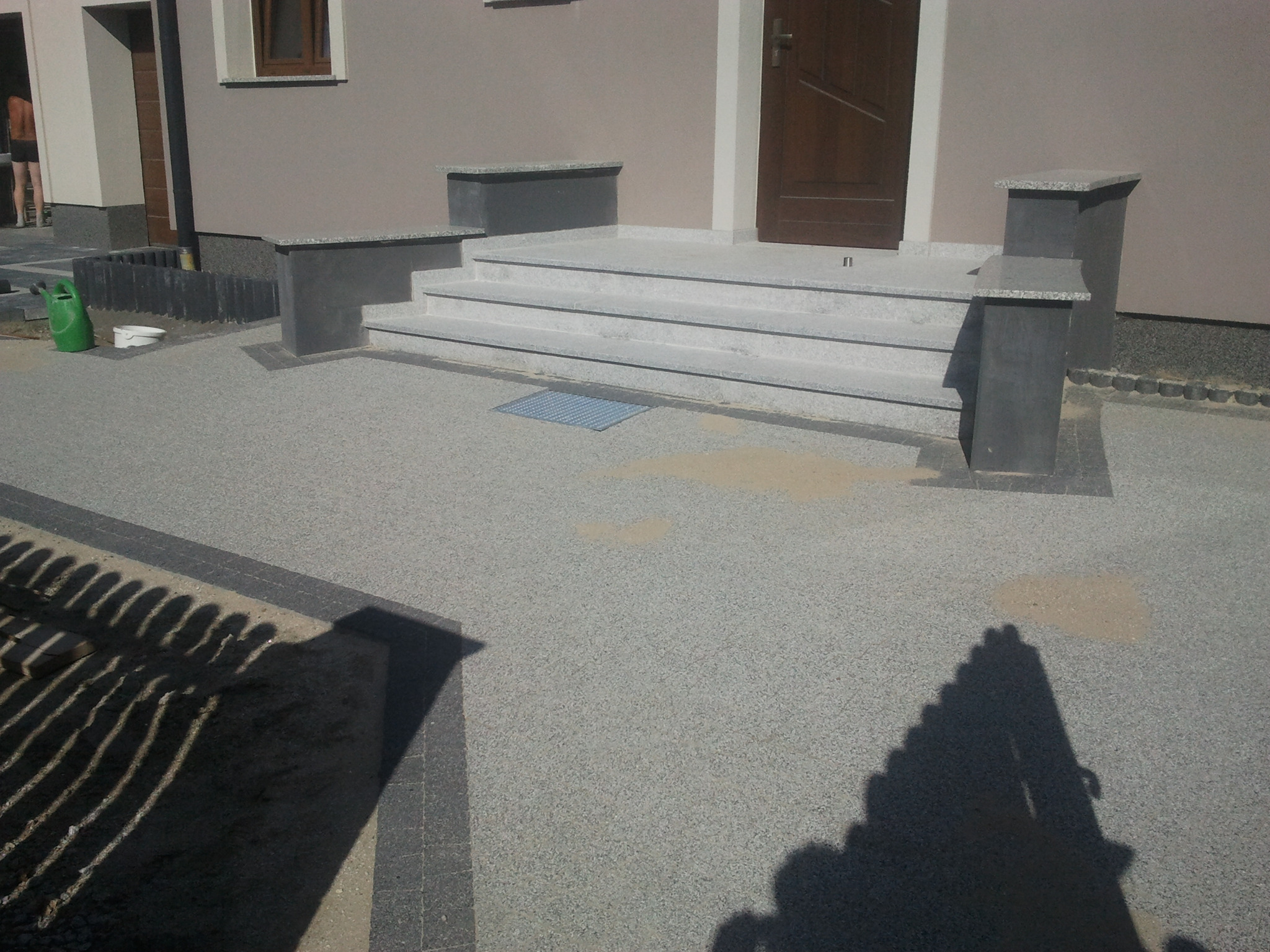 Nowe schody wejściowe z szarego kamienia prowadzące do domu, otoczone szarą nawierzchnią z drobnymi kamyczkami, widoczne cienie ogrodzenia i osoby w tle.