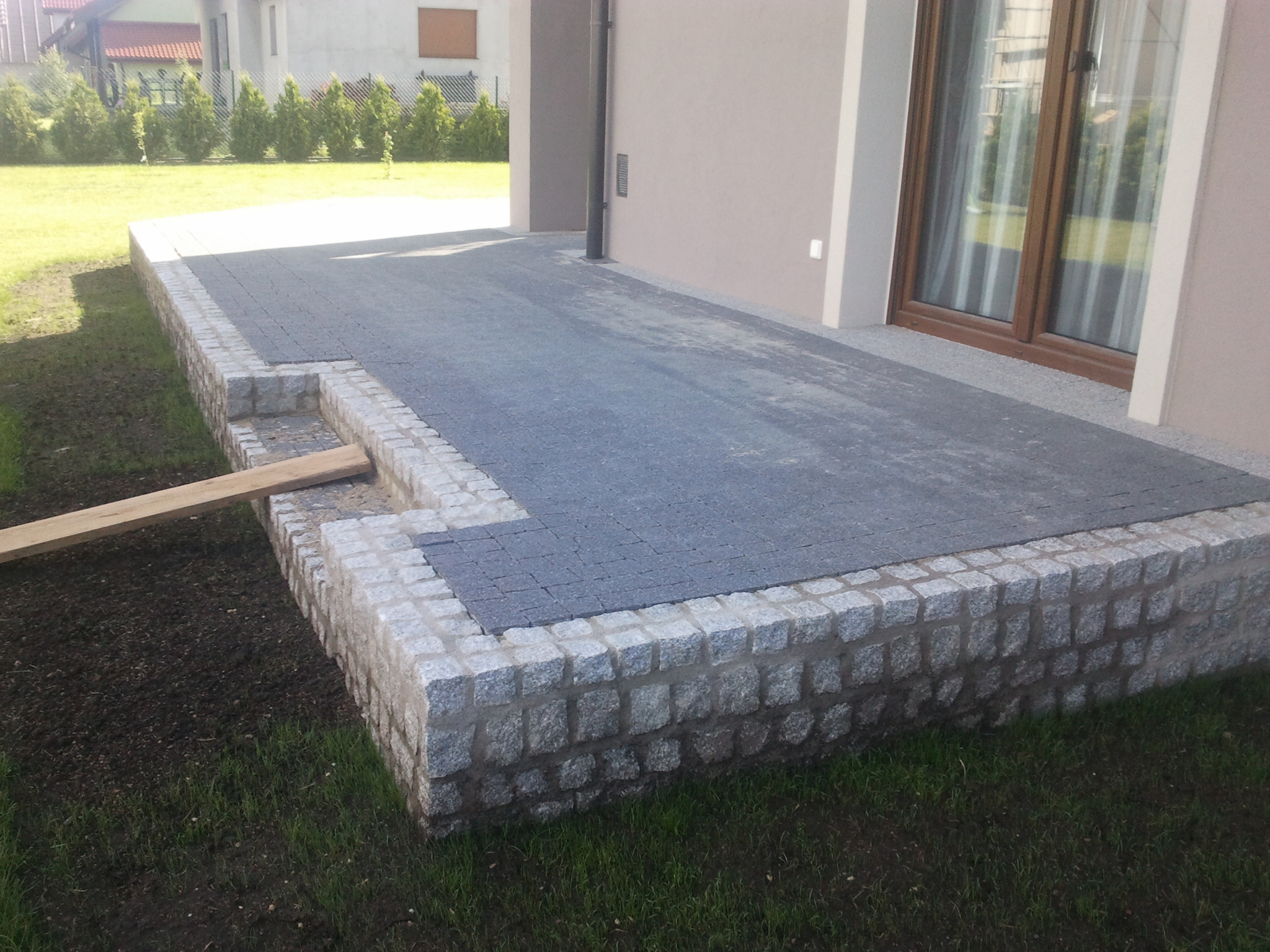 Granitowa obudowa tarasu z ciemnoszarą kostką brukową, widoczne schody z deski, trawnik i fragment domu w tle.