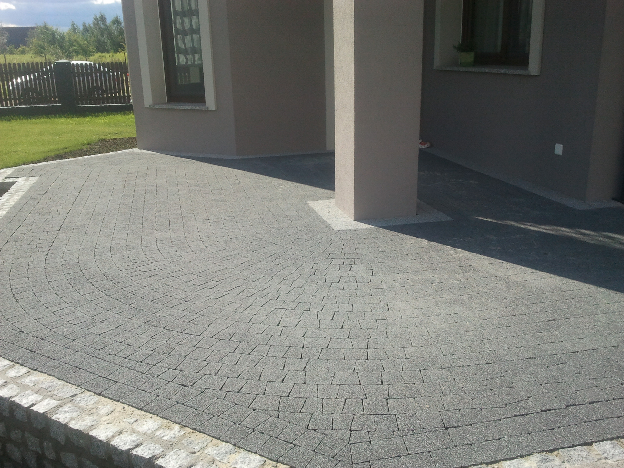 Półokrągły taras z szarej kostki brukowej ułożonej wachlarzowo, obrzeże z jasnych kamieni, kolumna budynku w tle.