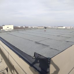 MUR-BETON MICHAŁ KLIMEK - Widok z góry na płaski dach pokryty papą termozgrzewalną na budynku wielorodzinnym, z widocznymi elementami obróbki blacharskiej oraz fragmentami rusztowania w kadrze, na tle panoramy miasta...