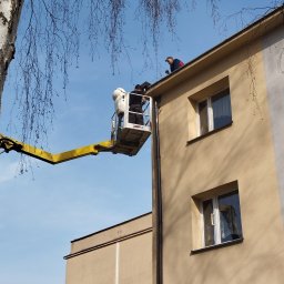 MUR-BETON MICHAŁ KLIMEK - Pracownicy na podnośniku koszowym przy budynku z beżową elewacją, jeden na dachu, drugi w koszu, prawdopodobnie wykonują prace dekarskie przy rynnach.