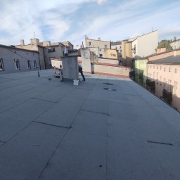 MUR-BETON MICHAŁ KLIMEK - Szary dach pokryty papą termozgrzewalną w otoczeniu kamienic, widoczna osoba pracująca na dachu i białe wiadro