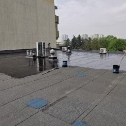 MUR-BETON MICHAŁ KLIMEK - Płaski dach pokryty papą, częściowo zabezpieczony czarną, błyszczącą powłoką bitumiczną. Widoczne puszki z preparatem, wałek malarski i pracownik w trakcie aplikacji. W tle budynek mieszkalny...