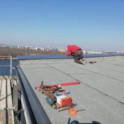 MUR-BETON MICHAŁ KLIMEK - Dekarz w czerwonej bluzie klęczy na płaskim dachu pokrytym papą, wykonując obróbki blacharskie, z widokiem na miasto i jezioro w tle, obok leżą narzędzia pracy.