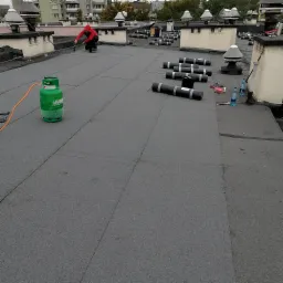 Prace dekarskie w Dąbrowie Górniczej: dekarz w czerwonej bluzie zgrzewa papę na płaskim dachu, obok leżą rolki papy i butla gazowa.