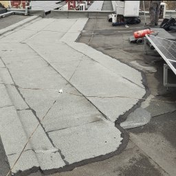 MUR-BETON MICHAŁ KLIMEK - Płaski dach pokryty papą bitumiczną w trakcie renowacji, widoczne świeże uszczelnienia i kable uziemiające, w tle panele słoneczne i sprzęt budowlany.