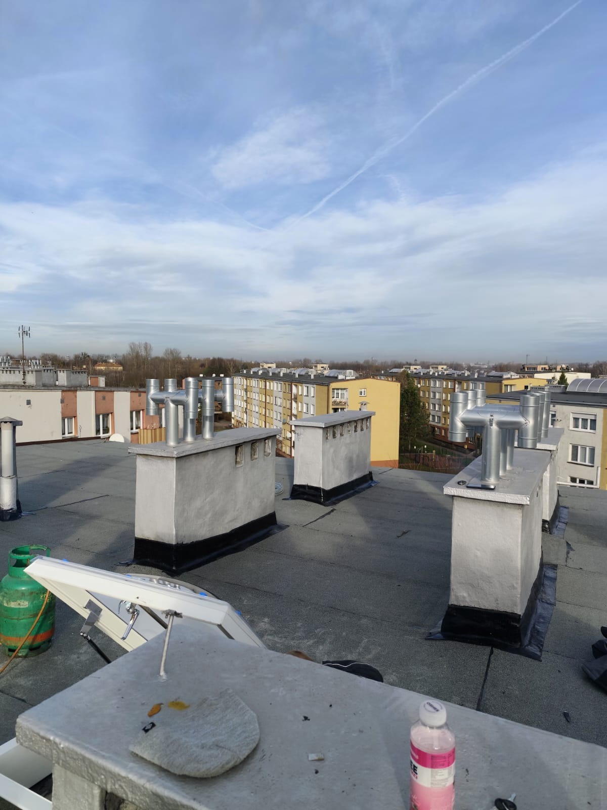 Widok z góry na płaski dach bloku z kilkoma kominami wentylacyjnymi z metalowymi rurami, butla gazowa i otwarte okno dachowe w trakcie montażu, na tle zabudowa miejska i błękitne niebo z chmurami.