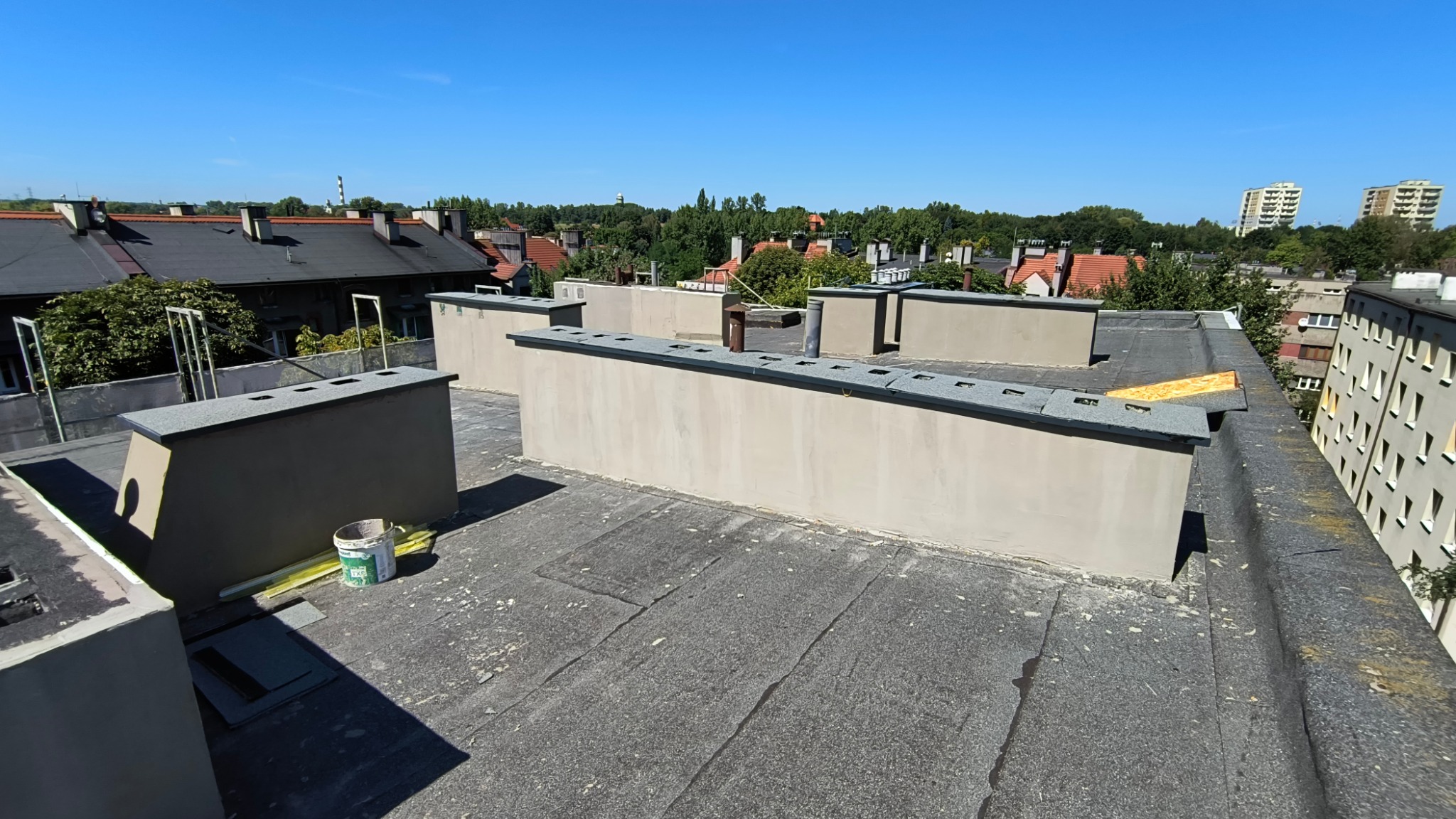 Widok z góry na płaski dach z wykończonymi attykami, widoczny kubeł z materiałami budowlanymi oraz fragmenty papy, w tle zabudowa miejska i błękitne niebo.