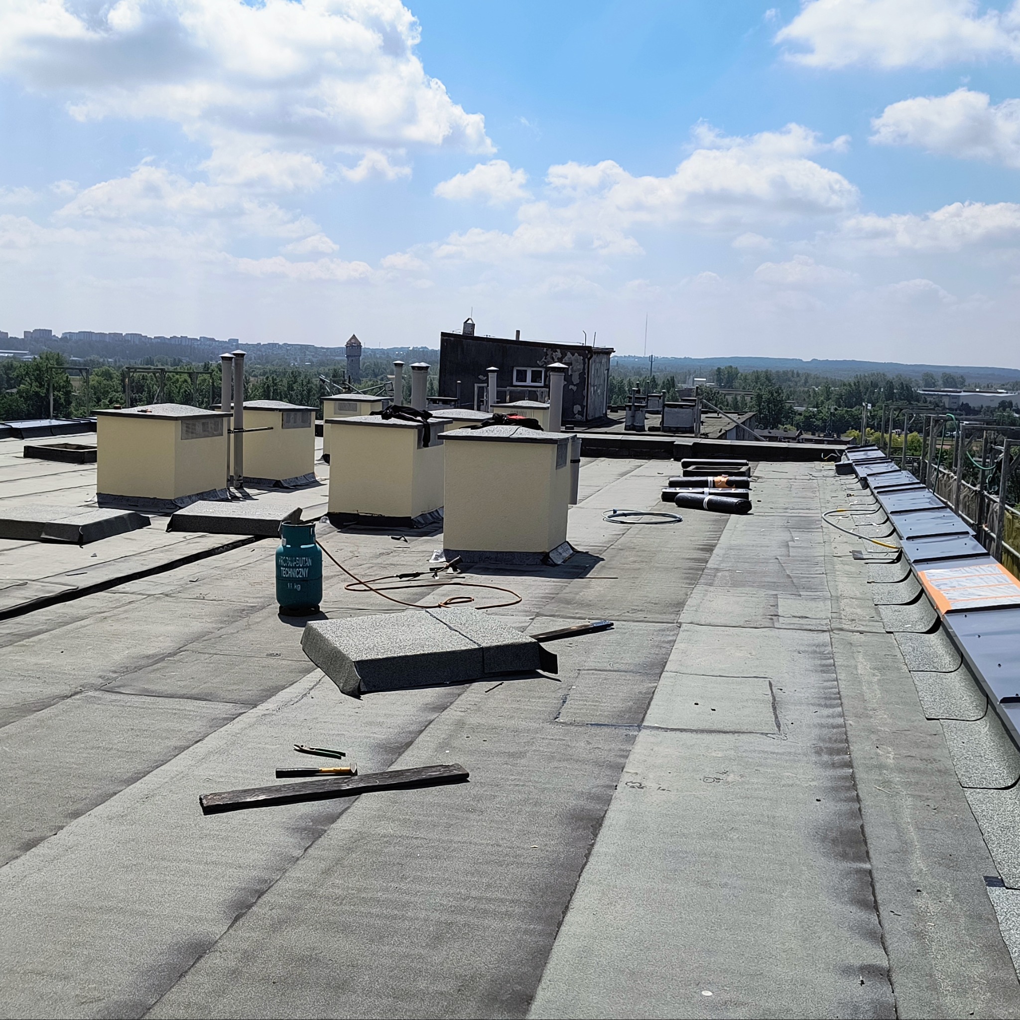 Widok z góry na płaski dach pokryty papą termozgrzewalną, z widocznymi kominami wentylacyjnymi, butlą gazową i narzędziami dekarskimi, w tle zabudowa miejska pod błękitnym niebem z chmurami.