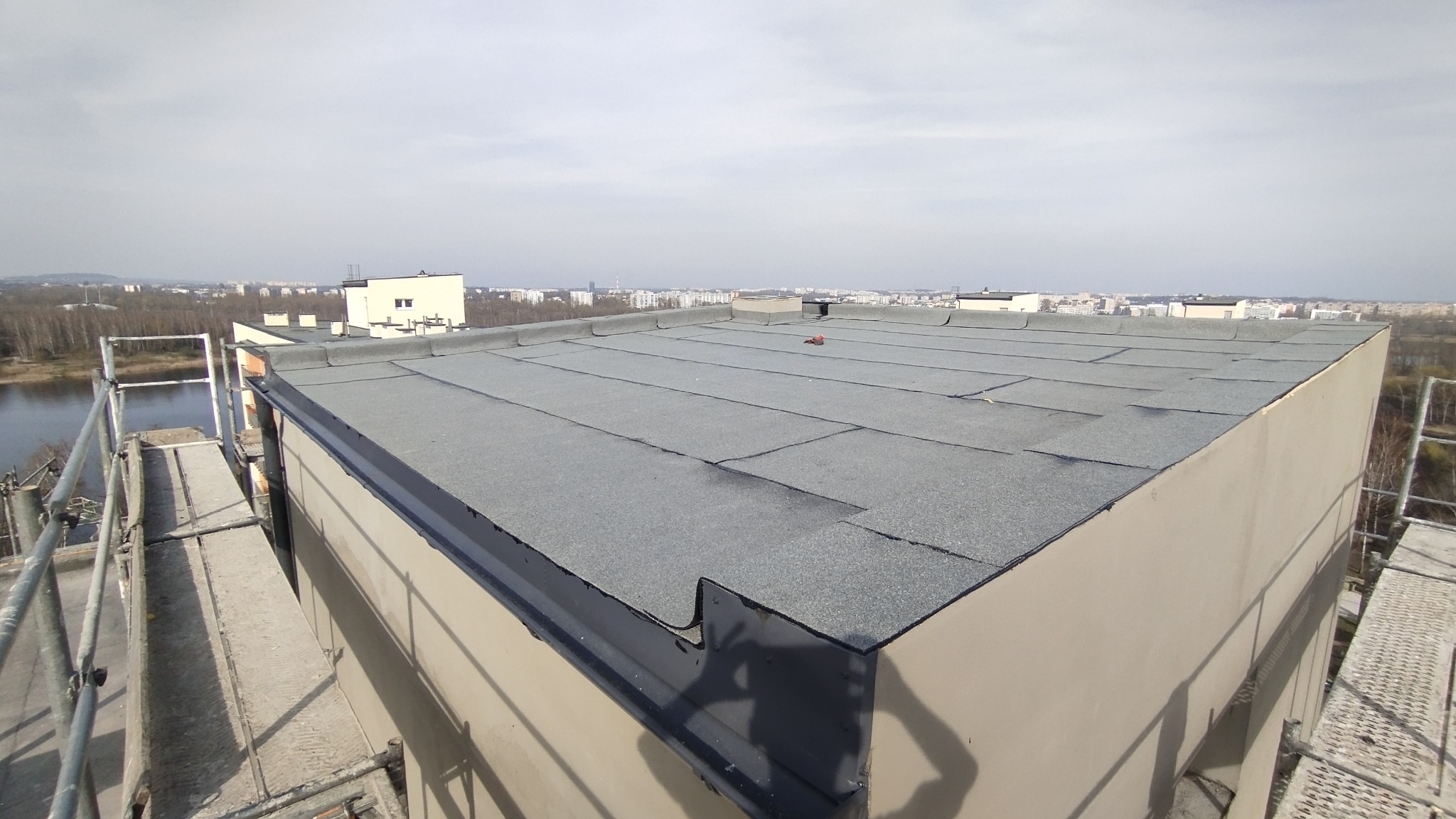 Widok z góry na płaski dach pokryty papą termozgrzewalną na budynku wielorodzinnym, z widocznymi elementami obróbki blacharskiej oraz fragmentami rusztowania w kadrze, na tle panoramy miasta...