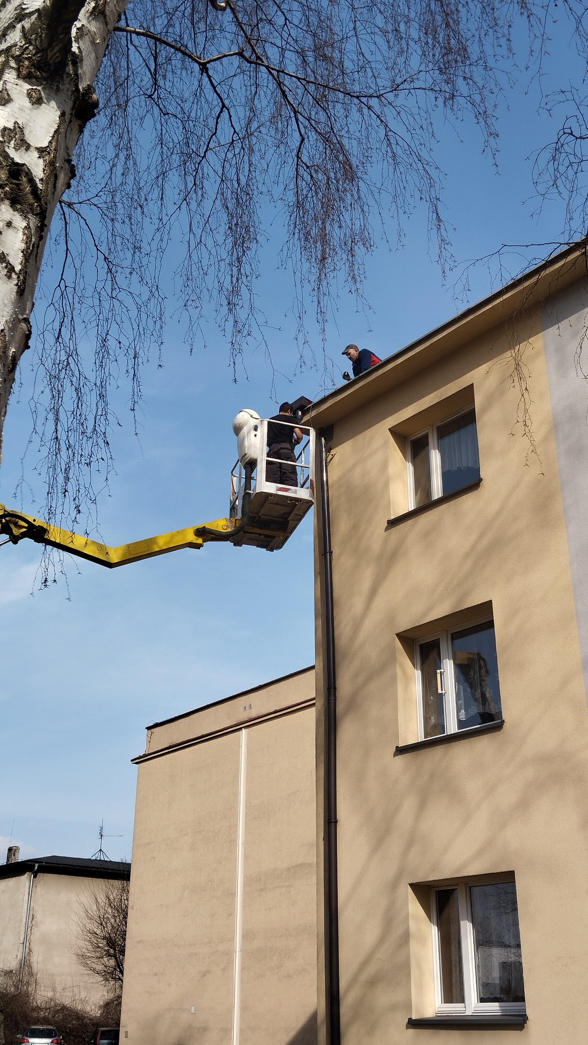 Pracownicy na podnośniku koszowym przy budynku z beżową elewacją, jeden na dachu, drugi w koszu, prawdopodobnie wykonują prace dekarskie przy rynnach.