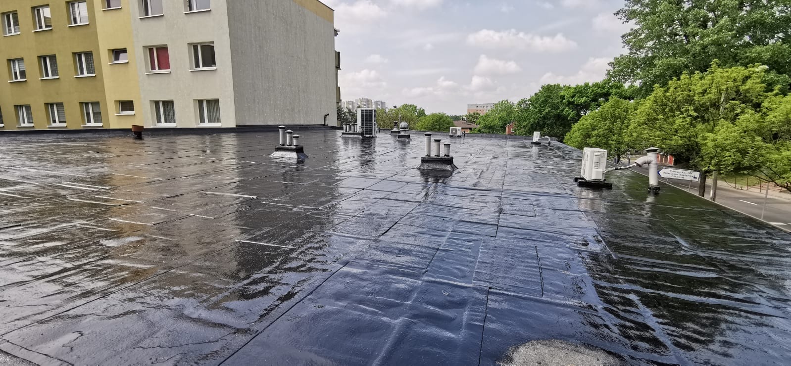 Widok z góry na płaski dach pokryty czarną, błyszczącą papą termozgrzewalną, z widocznymi kominami wentylacyjnymi i klimatyzatorami, na tle budynku mieszkalnego i zieleni drzew.
