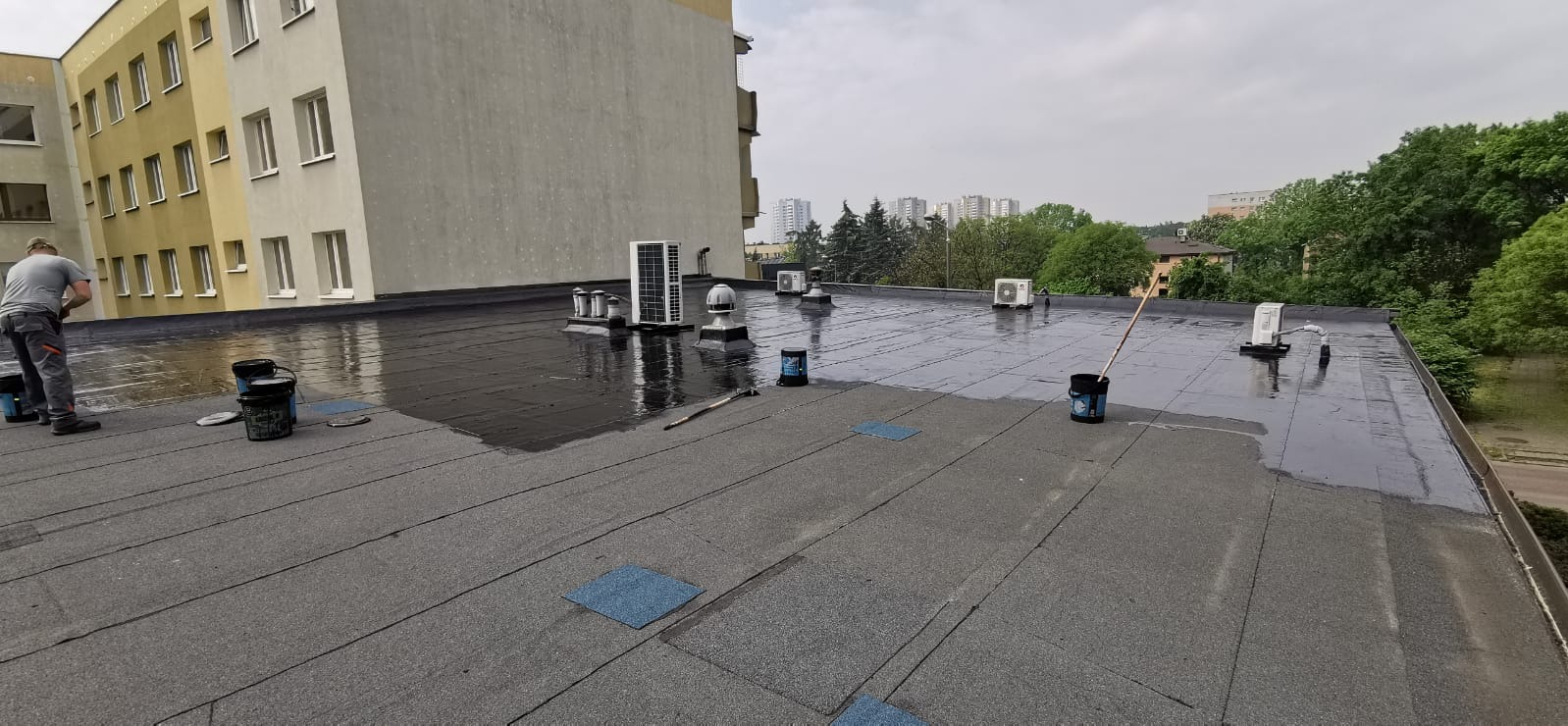 Płaski dach pokryty papą, częściowo zabezpieczony czarną, błyszczącą powłoką bitumiczną. Widoczne puszki z preparatem, wałek malarski i pracownik w trakcie aplikacji. W tle budynek mieszkalny...