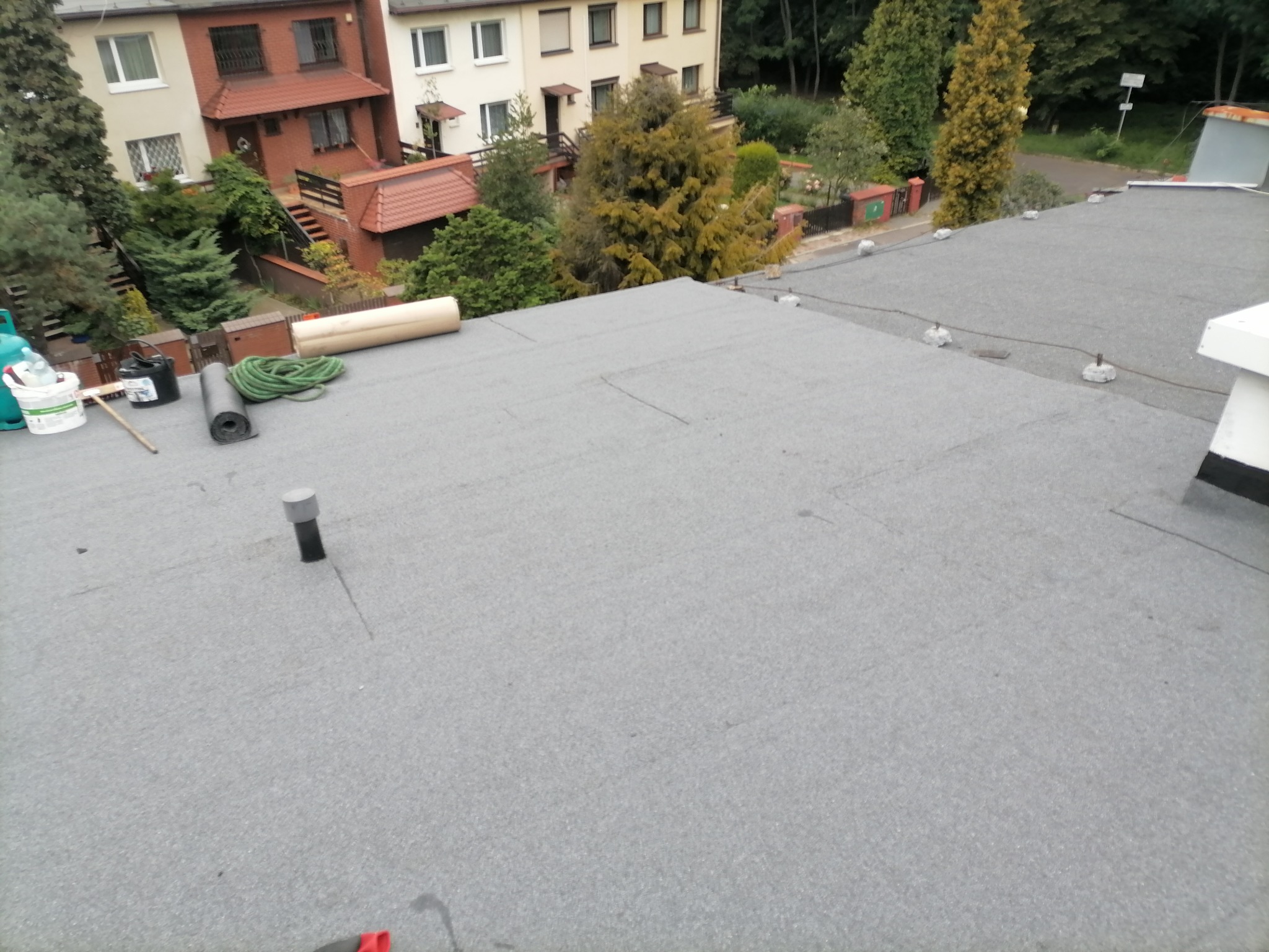 Szary dach pokryty papą termozgrzewalną z widocznymi elementami wentylacji, obciążnikami i narzędziami dekarskimi na tle zabudowy jednorodzinnej z zielenią.