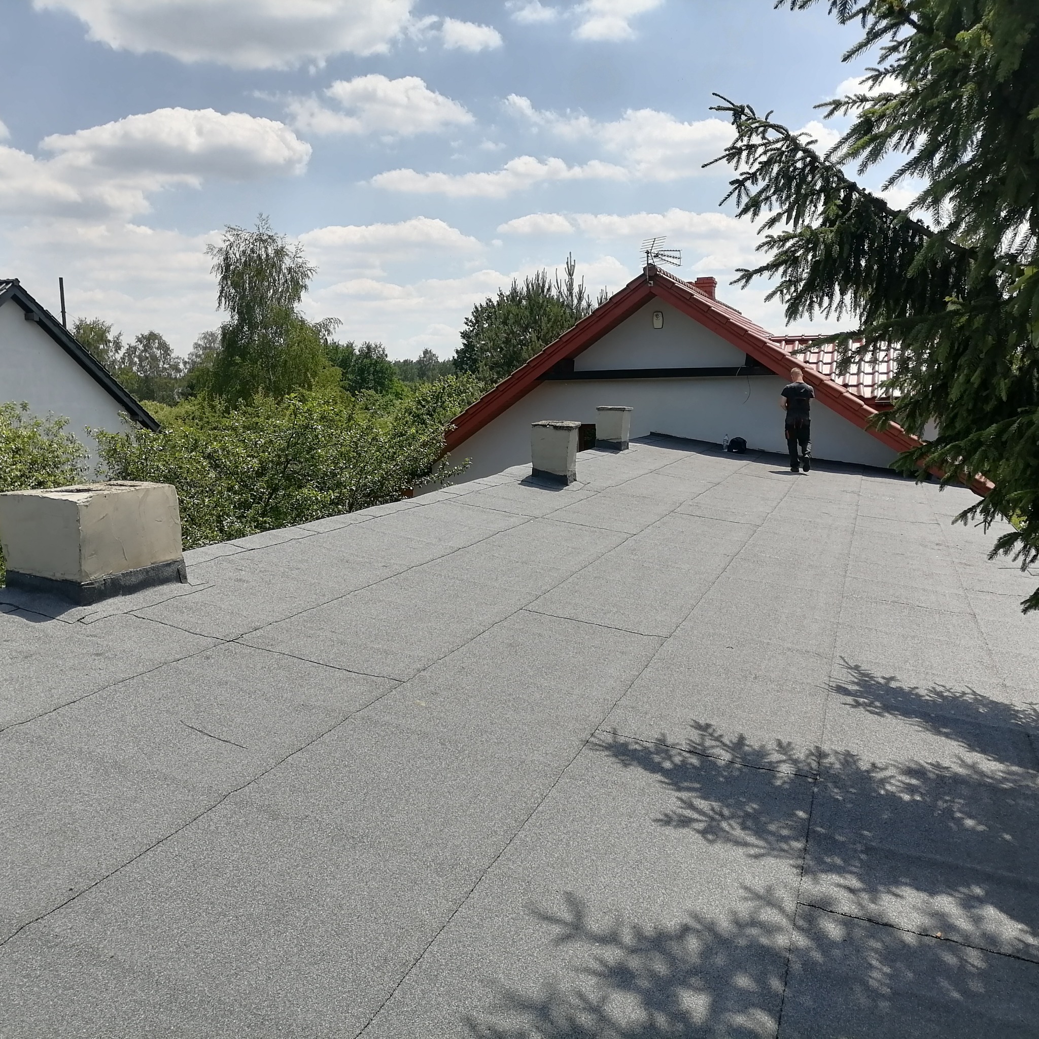 Szary, płaski dach pokryty papą termozgrzewalną z widocznymi kominami i sylwetką dekarza na tle błękitnego nieba z chmurami i zieleni drzew.