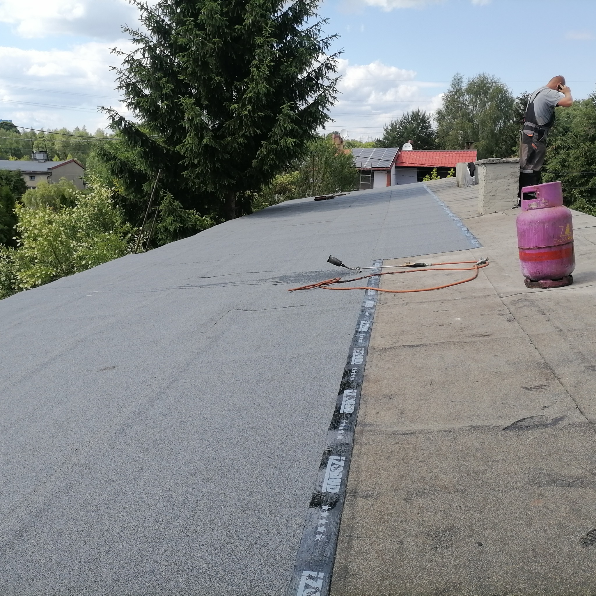 Pracownik w kombinezonie na dachu, obok różowa butla gazowa i palnik, widoczna nowa papa na połowie dachu i stara betonowa powierzchnia na drugiej połowie, w tle zabudowania i drzewa.