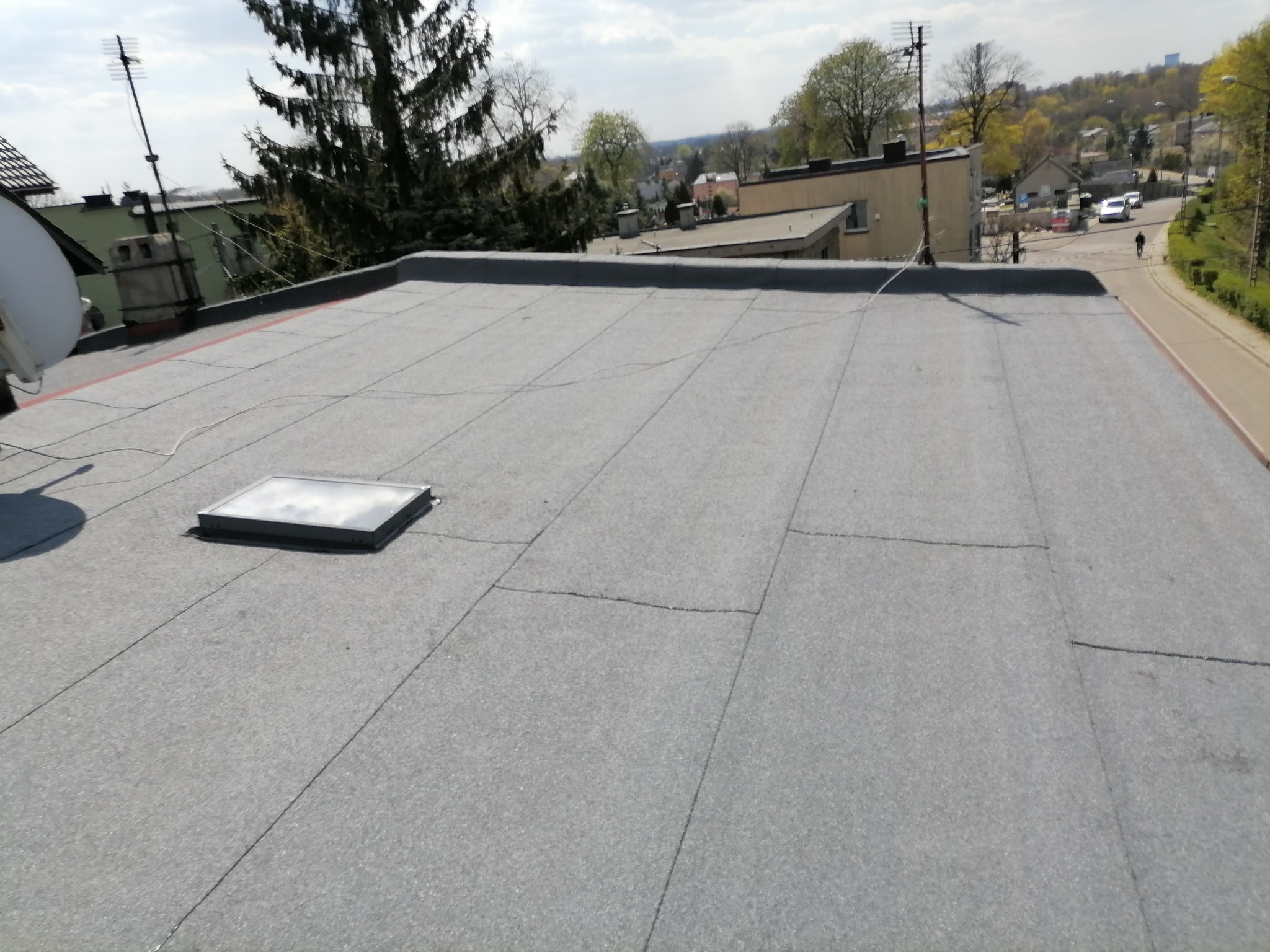 Szary, płaski dach pokryty papą z widocznym świetlikiem dachowym, anteną satelitarną i kominem w tle, z elementami zieleni i zabudowy miejskiej w oddali.