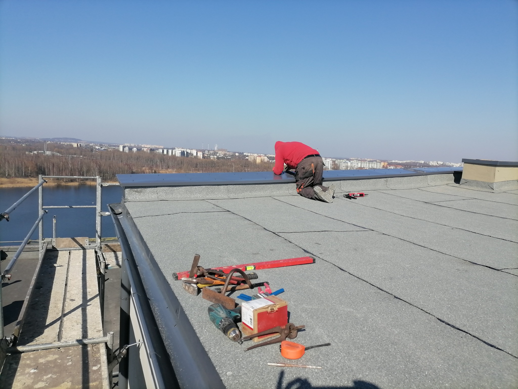 Dekarz w czerwonej bluzie klęczy na płaskim dachu pokrytym papą, wykonując obróbki blacharskie, z widokiem na miasto i jezioro w tle, obok leżą narzędzia pracy.