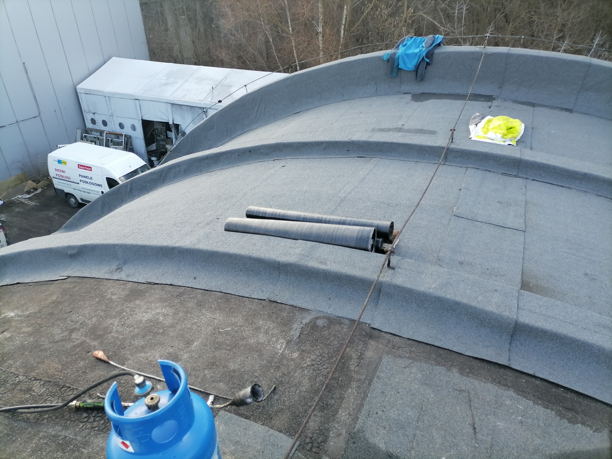 Widok z góry na płaski dach pokryty papą z widocznymi dwoma rurami wentylacyjnymi, butlą gazową, narzędziami dekarskimi oraz elementami odzieży roboczej. W tle budynek przemysłowy i biały namiot.