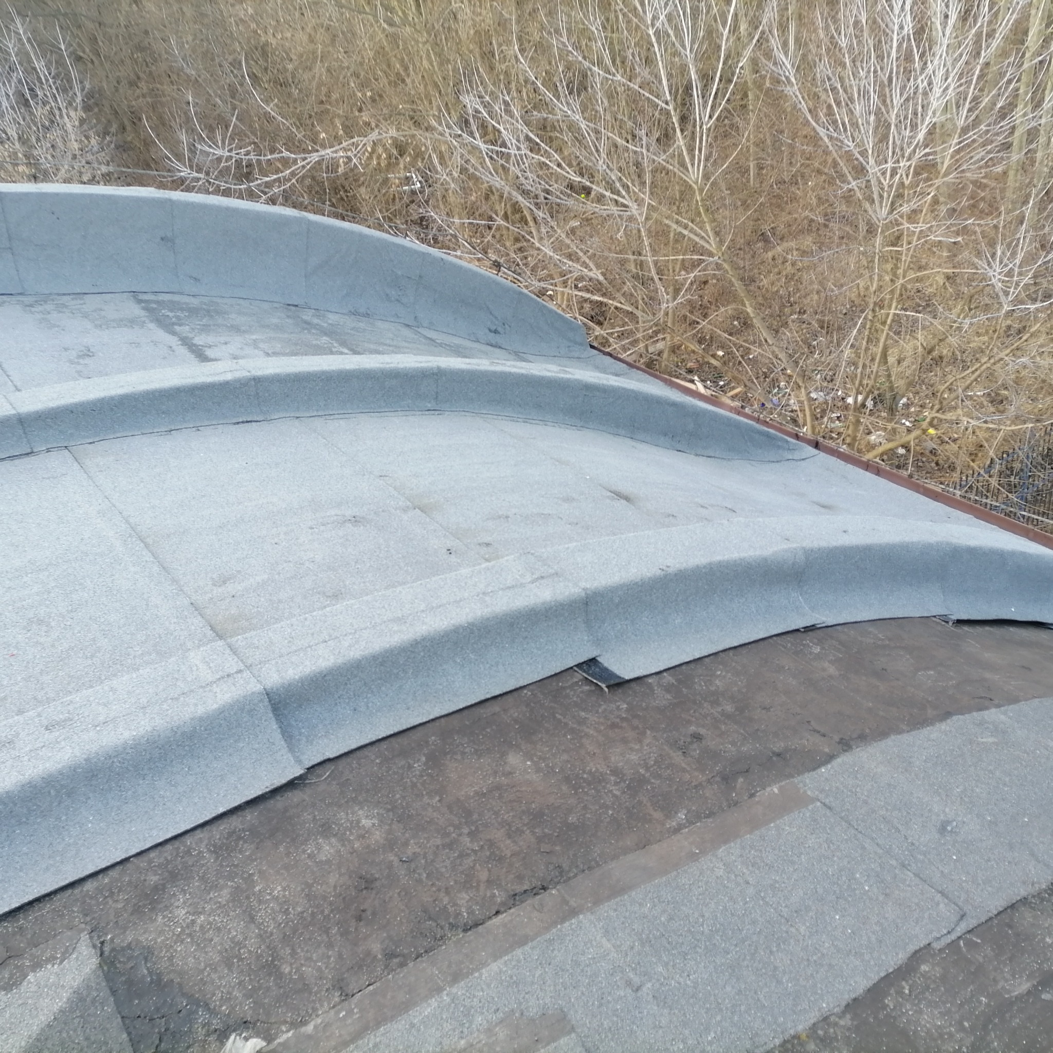 Fragment dachu pokrytego papą termozgrzewalną, z widocznymi łączeniami i elementami wykończeniowymi; w tle bezlistne krzewy