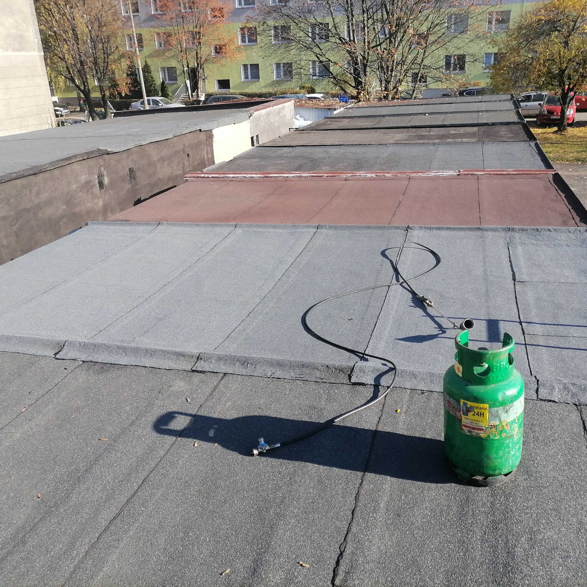 Butla gazowa z podłączonym palnikiem leży na płaskim dachu pokrytym papą termozgrzewalną, w tle budynek mieszkalny.